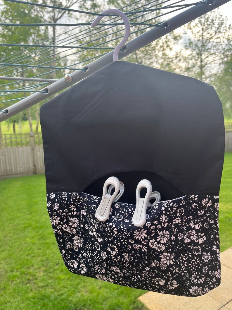 Puede incluir: Una bolsa de lavander&iacute;a de tela negra con estampado floral y un gancho blanco. La bolsa tiene un bolsillo en la parte delantera para guardar pinzas para la ropa. Hay tres pinzas para la ropa blancas en el bolsillo.