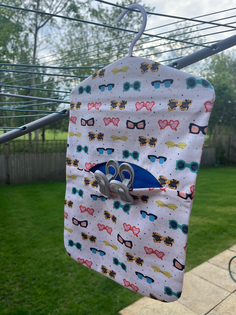Puede incluir: Un organizador colgante de tela blanca con una percha, que presenta un patr&oacute;n repetitivo de gafas de sol de colores y corazones. El organizador tiene un bolsillo azul con un juego de pinzas de ropa plateadas en el interior.