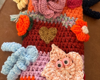 May include: A rectangular crochet sensory mat featuring a vibrant orange base with horizontal stripes of red, purple, and pink. It is decorated with a large pink flower, a gold heart, a peach-coloured star with googly eyes, and blue accents.