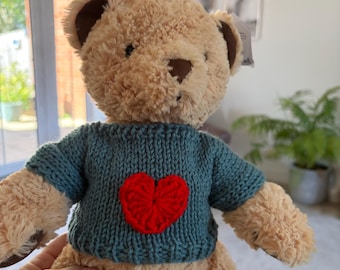 May include: A plush teddy bear with light brown fur, dark brown ears, and a dark brown nose. The bear is wearing a teal knitted sweater featuring a prominent, textured red heart stitched onto the centre of the chest area.