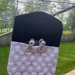 Puede incluir: Una bolsa de lavander&iacute;a de tela negra con un patr&oacute;n de sol y luna blanco y gris. La bolsa tiene un gancho para colgar y dos pinzas para la ropa sujetas a la parte superior.