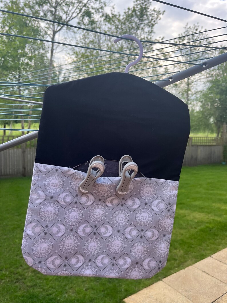 Puede incluir: Una bolsa de lavander&iacute;a de tela negra con un patr&oacute;n de sol y luna blanco y gris. La bolsa tiene un gancho para colgar y dos pinzas para la ropa sujetas a la parte superior.