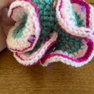 May include: A handcrafted crochet flower in pink, white, and teal, adorned with purple beads. The flower features layered petals with a dark pink border. The item is held against a wooden background.