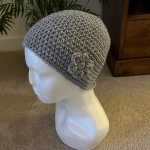 Pode incluir: Um gorro de crochê cinza-claro com um pequeno detalhe de flor de crochê a condizer na lateral. O gorro é feito com um padrão de pontos texturizado e consistente e é exibido num manequim de cabeça branco liso.