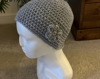 Puede incluir: Un gorro de ganchillo gris claro que presenta un pequeño adorno de flor de ganchillo a juego en el lateral. El gorro está confeccionado con un patrón de punto texturizado y consistente, y se muestra sobre un maniquí de cabeza blanco.