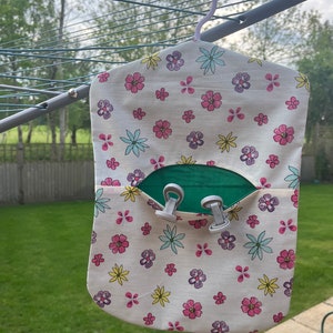 Puede incluir: Una bolsa de lavander&iacute;a de tela blanca con estampado floral en rosa, amarillo, azul y morado. La bolsa tiene un bolsillo verde con dos clips blancos. La bolsa est&aacute; colgada de un tendedero.