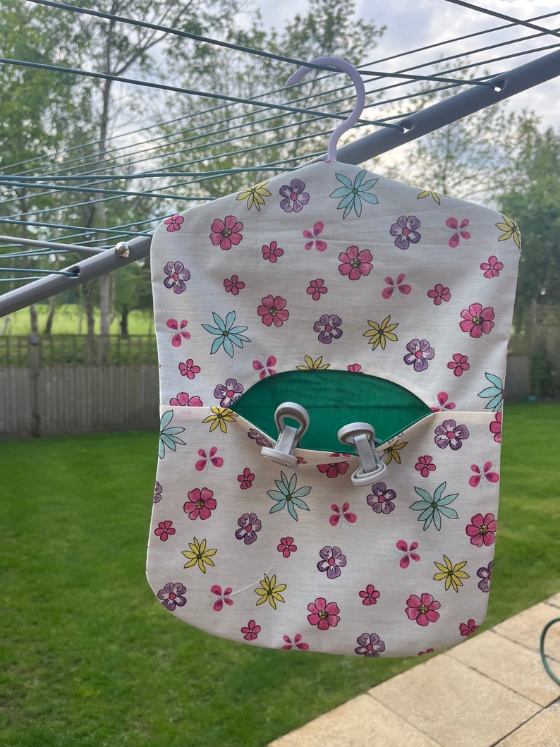 Puede incluir: Una bolsa de lavander&iacute;a de tela blanca con estampado floral en rosa, amarillo, azul y morado. La bolsa tiene un bolsillo verde con dos clips blancos. La bolsa est&aacute; colgada de un tendedero.
