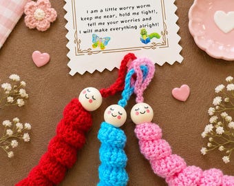 May include: Three crochet worry worms in red, blue, and pink, each with a wooden bead head featuring a painted face. A white card with scalloped edges displays the text The Worry Worm Poem and includes illustrations of a green worm and a butterfly.