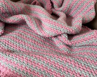 May include: A handmade crochet blanket featuring alternating horizontal stripes of soft pink and light grey yarn. The textile displays a consistent, textured stitch pattern throughout, creating a dense and uniform appearance across the fabric.