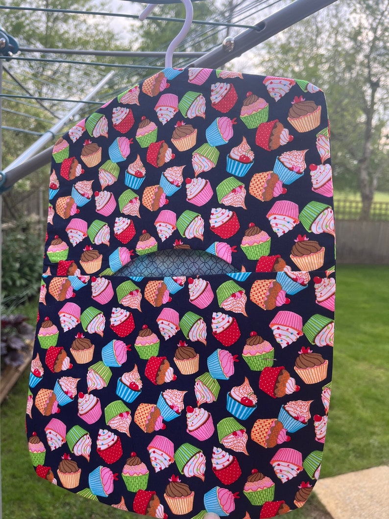 Pode incluir: Um cabide de tecido azul com um bolso na frente. O cabide &eacute; decorado com um padr&atilde;o repetido de cupcakes coloridos.