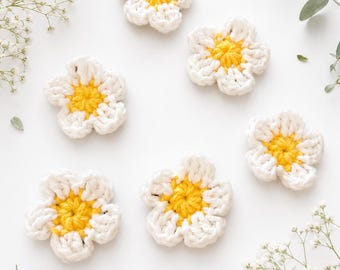 May include: Five individual crochet flowers, each featuring five white petals surrounding a bright yellow centre. The flowers are arranged in a scattered pattern, showcasing the textured yarn stitches and the consistent design of each handmade piece.