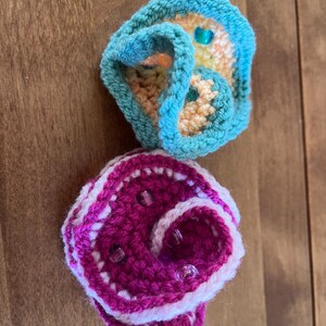 May include: Two handmade crochet items. The top item is teal, yellow, and orange with a teal border and a blue bead. The bottom item is pink and white with clear beads. Both items are on a wooden surface.