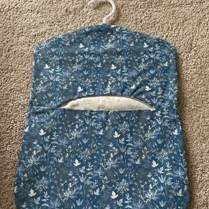 May include: A blue fabric garment bag with a white floral pattern. The bag has a white hanger and a pocket on the front.