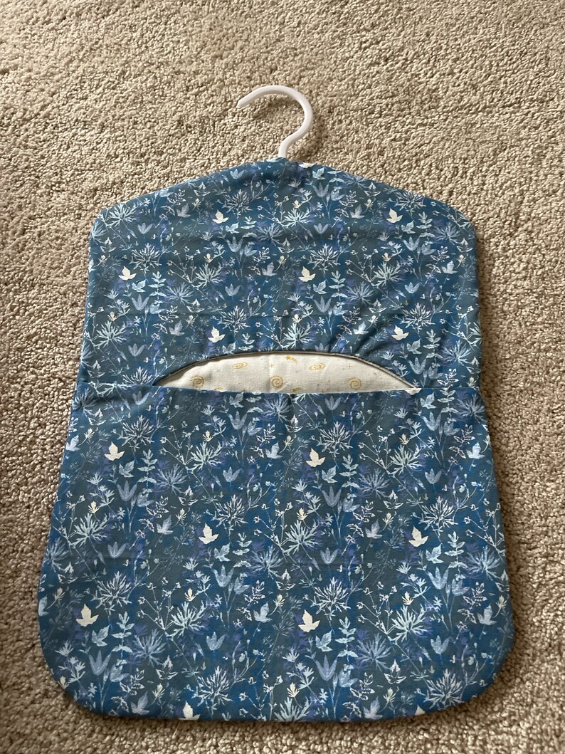 May include: A blue fabric garment bag with a white floral pattern. The bag has a white hanger and a pocket on the front.