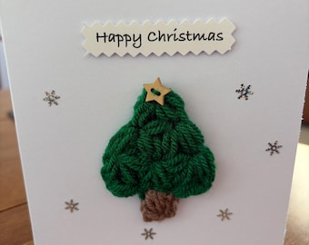 May include: A white greeting card featuring a crocheted green Christmas tree with a brown trunk and a small wooden star button at the top. The text Happy Christmas is printed on a white scalloped paper strip above the tree, surrounded by silver snowflakes.