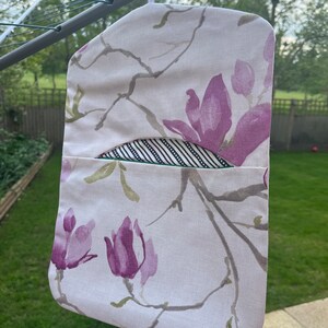 Pode incluir: Uma bolsa de roupa de tecido branco com estampa floral de flores de magn&oacute;lia rosa e galhos verdes. A bolsa tem um forro de tecido listrado preto e branco e um la&ccedil;o para pendurar.