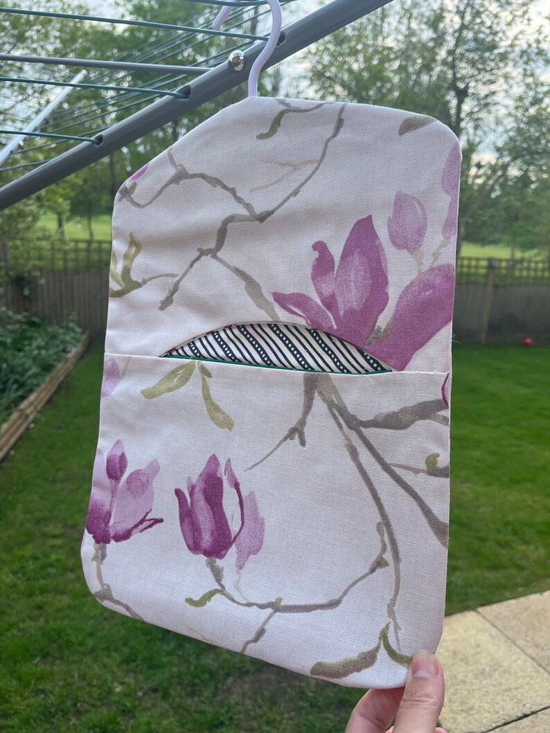 Pode incluir: Uma bolsa de roupa de tecido branco com estampa floral de flores de magn&oacute;lia rosa e galhos verdes. A bolsa tem um forro de tecido listrado preto e branco e um la&ccedil;o para pendurar.