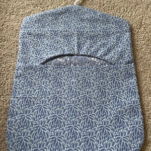 May include: A blue and white floral patterned garment bag with a white plastic hanger. The bag has a flap that opens to reveal a pocket for storing items.