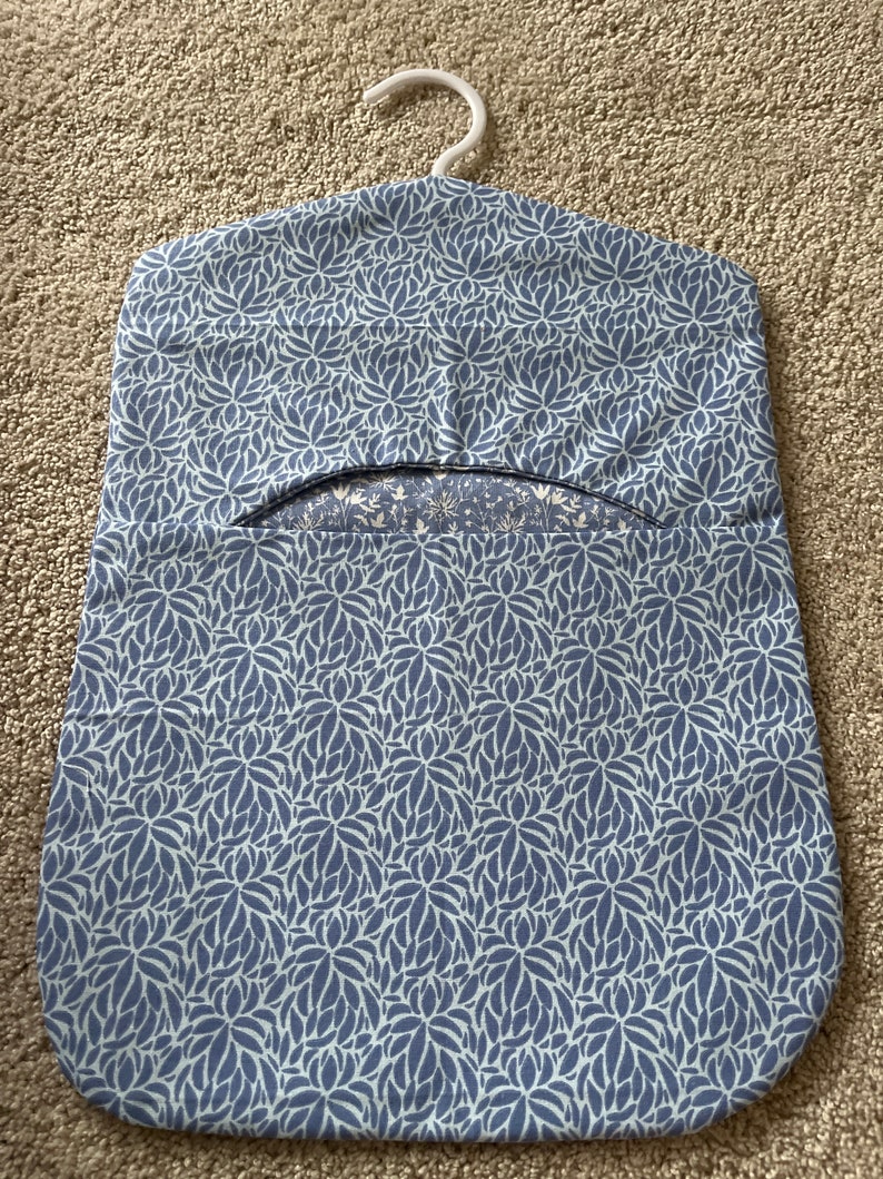 May include: A blue and white floral patterned garment bag with a white plastic hanger. The bag has a flap that opens to reveal a pocket for storing items.