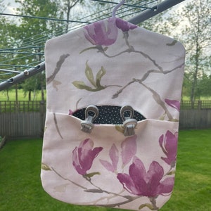 Puede incluir: Una bolsa de lavander&iacute;a de tela blanca con un estampado floral de flores de magnolia rosas y hojas verdes. La bolsa tiene una percha gris y un bolsillo con dos clips grises.