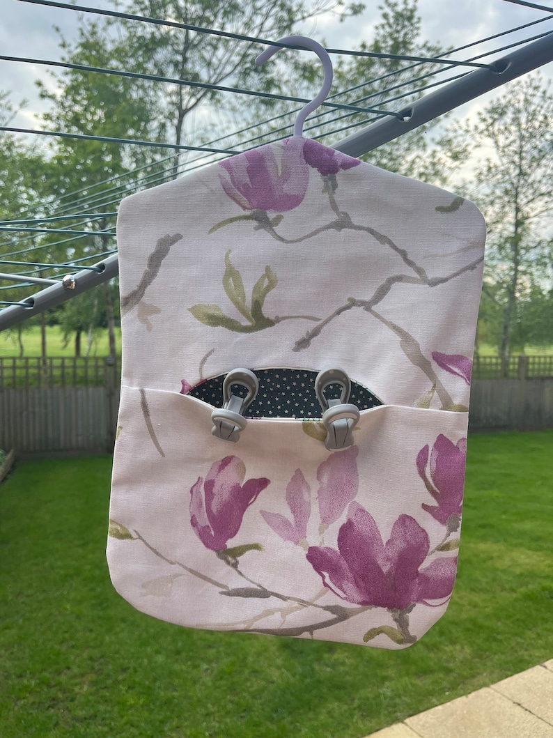 Puede incluir: Una bolsa de lavander&iacute;a de tela blanca con un estampado floral de flores de magnolia rosas y hojas verdes. La bolsa tiene una percha gris y un bolsillo con dos clips grises.