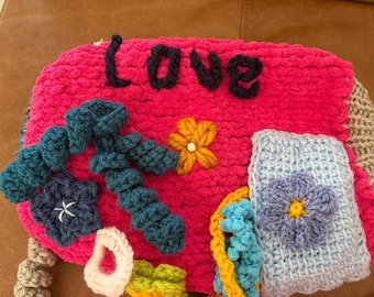 May include: A bright pink crocheted bag featuring the word Love in dark blue yarn. The bag is decorated with various crocheted elements including a yellow flower, a light blue rectangular patch with a purple flower, and dark blue curly accents.