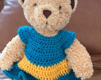 May include: A plush teddy bear with light tan, fuzzy fur and dark brown inner ears and nose. The bear is wearing a handmade, crocheted dress featuring a teal bodice and hem with a horizontal yellow stripe across the middle of the skirt.