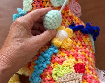 May include: A multi-coloured crochet sleeve featuring a variegated yarn base in shades of pink, orange, and yellow. The surface is decorated with various three-dimensional crochet appliqués, including light blue flowers, a yellow flower, and an ice cream cone.