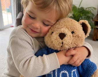 Puede incluir: Un oso de peluche color canela con ojos y nariz de color marrón oscuro, que lleva un suéter de punto azul con el nombre Allan bordado en hilo blanco en el pecho. El oso tiene un pelaje suave y texturizado y se sostiene en un abrazo.