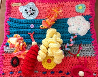 May include: A square crochet sensory mat featuring horizontal stripes in pink, teal, and grey. It includes various attached tactile elements: a light blue cat face, a yellow spiral, a white flower, a red textured cylinder, and a red heart with a bead.