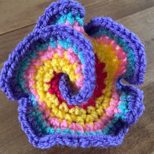 May include: A crocheted flower with a purple, pink, yellow and turquoise colour scheme. The flower is made with a spiral pattern and has a ruffled edge.