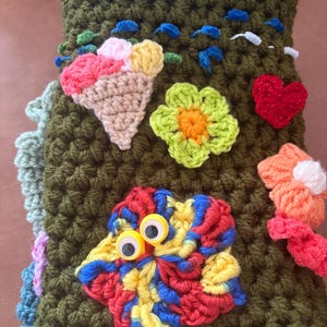 Pode incluir: Uma capa de crochê verde-oliva com vários apliques coloridos, incluindo um cone de sorvete bege, uma flor verde-limão, um coração vermelho, uma flor laranja e uma flor multicolorida com dois olhos móveis de plástico.