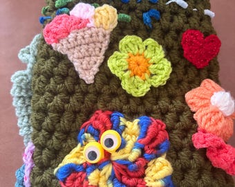 Puede incluir: Una funda de ganchillo verde oliva con varios apliques coloridos, incluyendo un cono de helado beige, una flor verde lima, un corazón rojo, una flor naranja y una flor multicolor con dos ojos móviles de plástico pegados.
