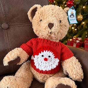 Pode incluir: Um urso de peluche bege com almofadas nas patas e nariz castanho escuro. O urso veste uma camisola de malha vermelha com um desenho de cara de Pai Natal em crochet branco na parte da frente. Uma etiqueta azul Jellycat está presa à orelha.