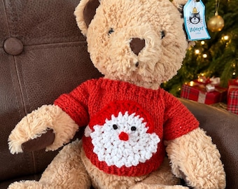 May include: A plush beige teddy bear with dark brown paw pads and a dark brown nose. The bear wears a red knitted jumper featuring a white crocheted Santa Claus face design on the front. A blue Jellycat brand tag is attached to the ear.