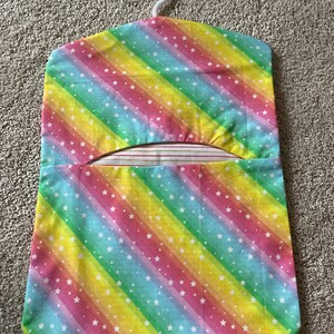 May include: A rainbow-coloured fabric hanger with white stars. The hanger has a white plastic hook and a pocket on the front.