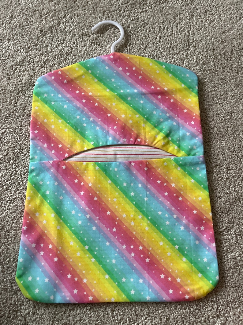 May include: A rainbow-coloured fabric hanger with white stars. The hanger has a white plastic hook and a pocket on the front.