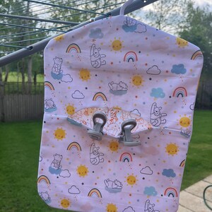 Puede incluir: Un organizador colgante de tela blanca con tem&aacute;tica de Winnie the Pooh. El organizador tiene un bolsillo en la parte delantera y dos clips en la parte superior. La tela presenta un patr&oacute;n repetitivo de personajes de Winnie the Pooh, arco&iacute;ris, nubes y estrellas.