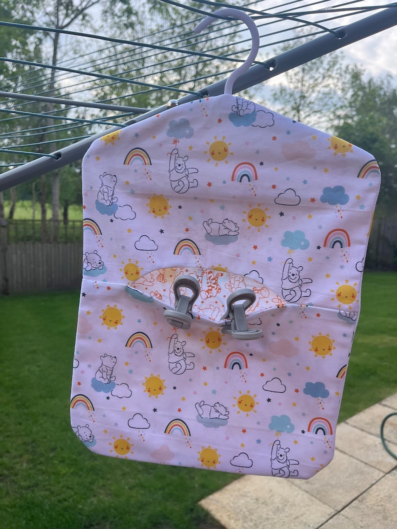Puede incluir: Un organizador colgante de tela blanca con tem&aacute;tica de Winnie the Pooh. El organizador tiene un bolsillo en la parte delantera y dos clips en la parte superior. La tela presenta un patr&oacute;n repetitivo de personajes de Winnie the Pooh, arco&iacute;ris, nubes y estrellas.