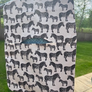 Pode incluir: Um organizador de pendurar de tecido branco com estampa de zebra em preto e branco. O organizador tem um bolso na frente e est&aacute; pendurado em um cabide.