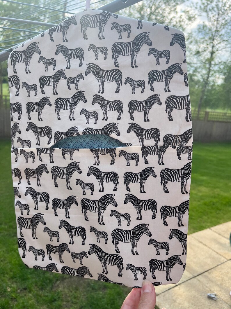 Pode incluir: Um organizador de pendurar de tecido branco com estampa de zebra em preto e branco. O organizador tem um bolso na frente e est&aacute; pendurado em um cabide.