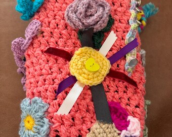 May include: A handcrafted crochet item in pink, blue, and purple, decorated with crochet flowers, ribbons, and a yellow button. The item has a textured surface and a whimsical design.