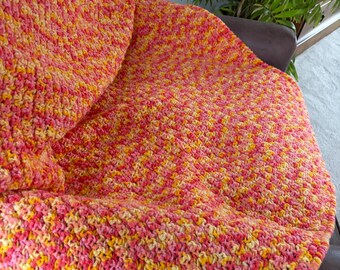 May include: A handmade crochet blanket featuring a variegated yarn pattern in vibrant shades of pink, orange, and yellow. The textile has a textured, looped stitch construction that creates a dense and consistent surface across the entire fabric.