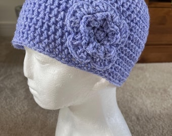 Puede incluir: Un gorro de ganchillo lila claro con un adorno floral. El gorro está hecho de hilo texturizado y presenta una flor decorativa en el lateral. El gorro se exhibe en una cabeza de maniquí blanca.