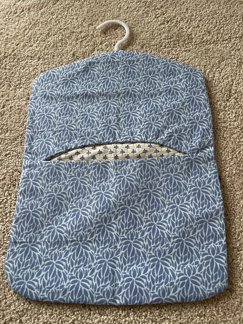 May include: A blue and white floral patterned laundry bag with a white plastic hanger. The bag has a small opening at the top for hanging clothes.