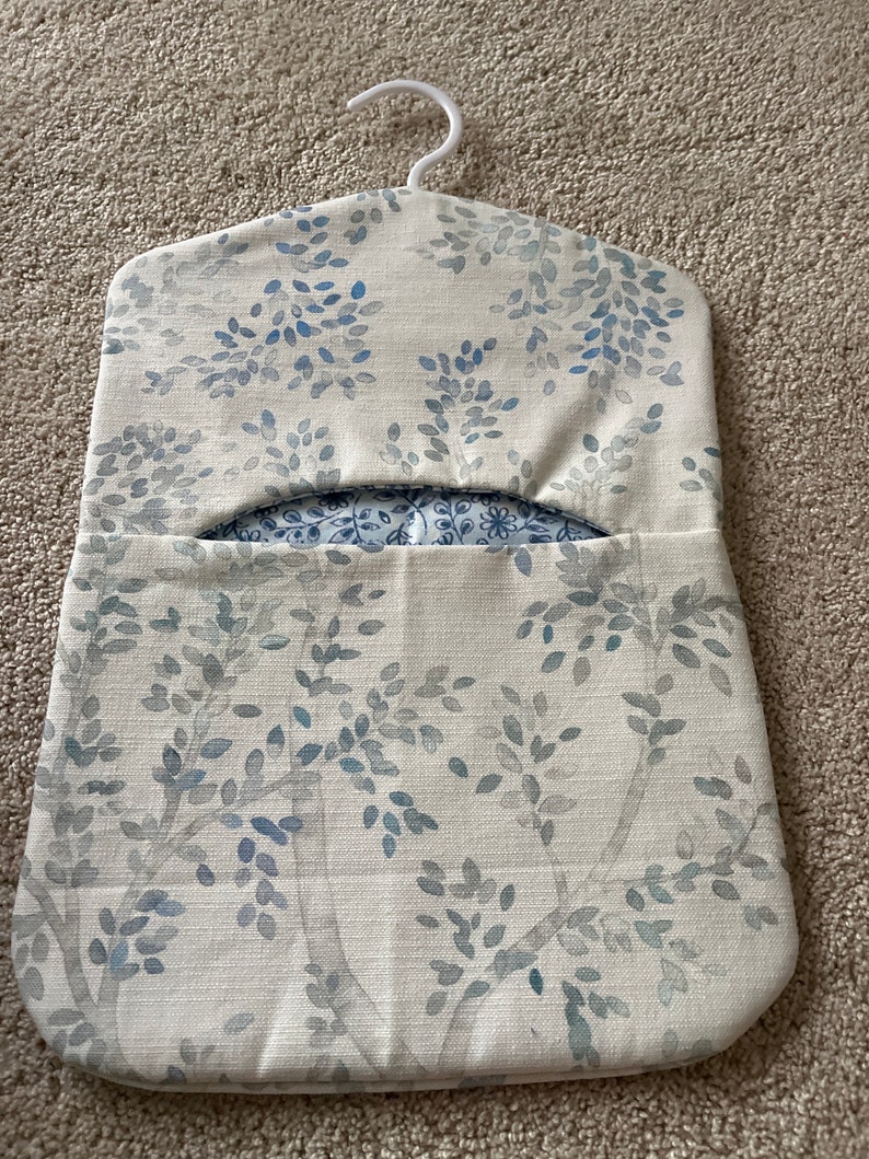 May include: A white fabric hanging organiser with a blue floral pattern. The organiser has a hanger at the top and a pocket in the front.