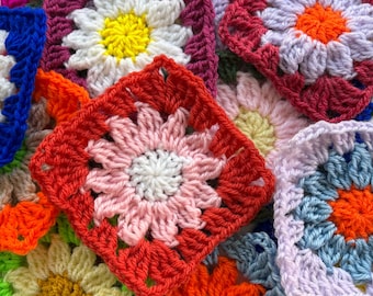 May include: A close-up of a pile of colourful crochet squares. Each square is a daisy design with petals in white, pink, orange, and yellow, with a contrasting border. The squares are in various colours, including red, blue, and green.