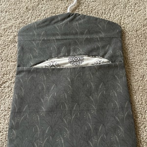 May include: A grey fabric hanging organiser with a white hanger. The fabric has a pattern of small grey leaves on a dark grey background. The organiser has a pocket on the front with a white fabric lining and a black and white geometric pattern.