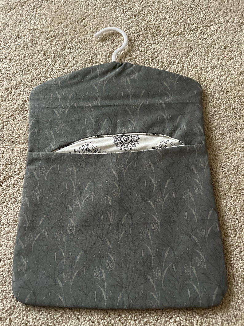 May include: A grey fabric hanging organiser with a white hanger. The fabric has a pattern of small grey leaves on a dark grey background. The organiser has a pocket on the front with a white fabric lining and a black and white geometric pattern.
