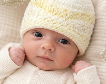 May include: A hand-crocheted baby beanie featuring a soft, variegated yarn pattern in shades of pale yellow and cream. The hat has a textured, repeating stitch design and a snug fit, crafted from a lightweight yarn suitable for a newborn infant.